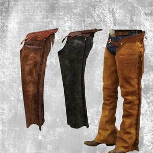 May include: Three pairs of leather chaps in shades of brown and black. The chaps have a western design, with decorative stitching and fringe. One pair is worn with denim jeans and cowboy boots, a classic look.