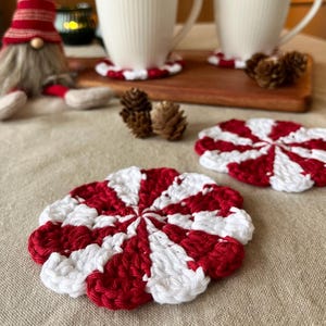 May include: Crocheted coasters in red and white, featuring a peppermint swirl pattern. The coasters are displayed on a neutral surface, with two white mugs and a wooden tray in the background. A Christmas gnome is also visible.