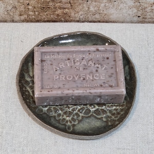 May include: A bar of artisanal soap on a ceramic soap dish. The soap is a light purple colour with flecks. The soap dish is mottled green and brown with a decorative, openwork design. The soap weighs 125g.