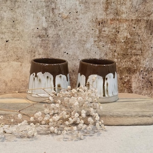 May include: Two ceramic cups featuring a glossy dark brown glaze on the upper portion that drips down over a matte white base. The cups have a wide, slightly tapered cylindrical shape with a smooth rim and a flat bottom, presented as a pair.