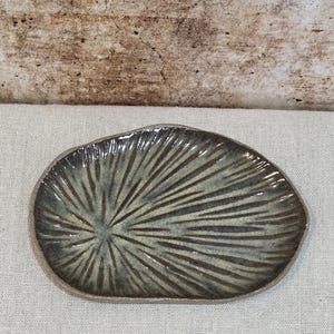 May include: A handmade ceramic dish with an organic shape. The dish has a radial pattern of dark brown lines against a muted green and grey background. The rim is a darker brown. The dish is approximately 15 cm in diameter.