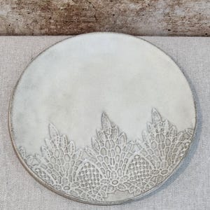 May include: A circular, cream-coloured ceramic plate featuring an embossed lace design along the lower edge. The plate has a textured finish and a muted, natural colour scheme, ideal for serving starters or desserts.