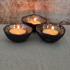 May include: Three black ceramic candle holders with lit candles. The holders have a dark, textured exterior and a reflective, golden interior. The candles are round and white, with a small flame.