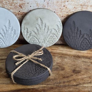 May include: Four round coasters in grey and black, featuring an embossed floral design. One stack is tied with twine. The coasters are displayed on a rustic wooden surface, with a variety of tones and textures.