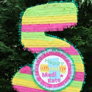 May include: A colorful, multi-layered, paper mache piñata shaped like the number five. The piñata is decorated with green, yellow, and pink tissue paper. A pink circle with white and green trim is attached to the front of the piñata. The circle has the text "Happy Birthday Madi Kate" in colorful letters.