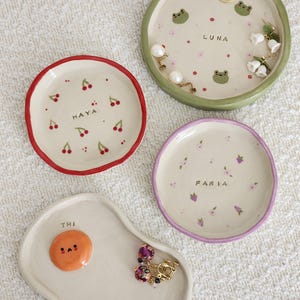 Puede incluir: Cuatro platos de cerámica para baratijas con diseños pintados. Uno tiene forma de huevo con una cara naranja, otro tiene cerezas y el nombre "HAYA", un tercero tiene ranas y el nombre "LUNA", y el último tiene flores y el nombre "FARIA".