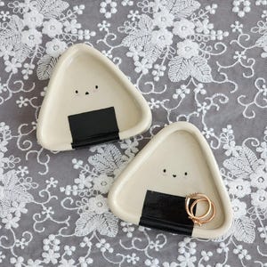 Puede incluir: Dos pequeños platos de cerámica color crema con forma de onigiri, con detalles de nori negro y pequeñas caras. Un plato contiene dos anillos dorados. Los platos están sobre una superficie de encaje blanco.