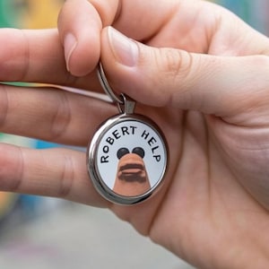 Könnte beinhalten: Ein silberner Schlüsselanhänger mit einem runden Anhänger, der ein Cartoon-Gesicht und den Text "ROBERT HELP" zeigt. Der Anhänger hat einen weißen Hintergrund, schwarze Ohren und eine braune Nase. Der Schlüsselanhänger wird in einer Hand gehalten.