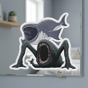 May include: A sticker depicting a dark grey monster with a large mouth and sharp teeth, and long, spindly limbs. Above it is a grey fish with an open mouth. The sticker is on a mirror.