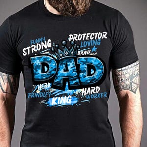 May include: A black short-sleeved t-shirt featuring the word DAD in large blue camouflage letters with a crown graphic on top. Surrounding the text are the words FUNNY, STRONG, PROTECTOR, LOVING, BRAVE, HERO, FRIENDLY, HARD WORKER, and KING.