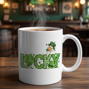 May include: White ceramic mug with a St. Patrick's Day theme. The mug features the word "LUCKY" in green with shamrock and plaid patterns, and a cartoon goose wearing a green hat. The mug is filled with hot coffee.