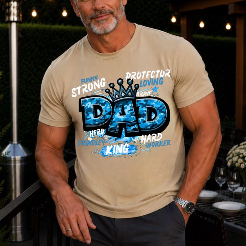May include: A beige short-sleeved t-shirt featuring a graphic design with the word DAD in large blue camouflage letters topped with a black crown. Surrounding text includes the words FUNNY, STRONG, PROTECTOR, LOVING, BRAVE, HERO, HARD WORKER, and KING.