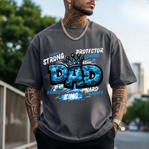 May include: A charcoal grey short-sleeve t-shirt featuring a large blue camouflage-patterned graphic that reads DAD with a crown icon above it. Surrounding text includes the words STRONG, PROTECTOR, LOVING, BRAVE, HERO, FRINDLY, and HARD WORKER.