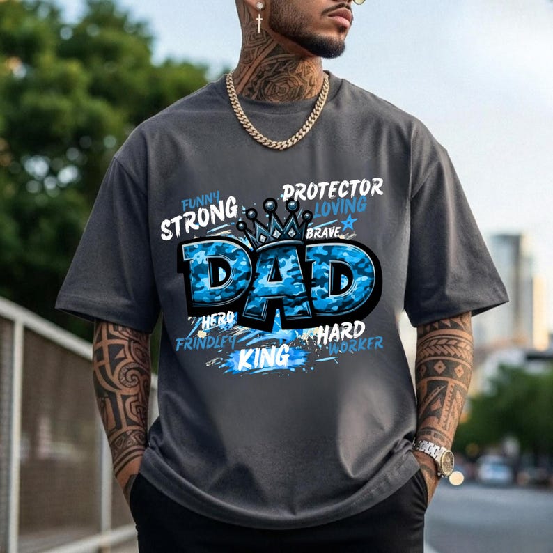 May include: A charcoal grey short-sleeve t-shirt featuring a large blue camouflage-patterned graphic that reads DAD with a crown icon above it. Surrounding text includes the words STRONG, PROTECTOR, LOVING, BRAVE, HERO, FRINDLY, and HARD WORKER.