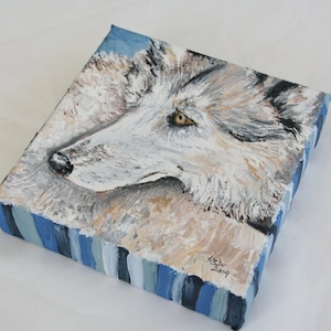 May include: A close-up of a painting of a wolf's head. The wolf is painted in shades of white, gray, and black, with a hint of blue in the background. The wolf's eye is a bright yellow. The painting is signed "K.S. 2019" in the lower right corner.