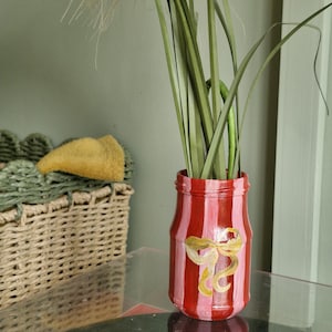 May include: A red and pink striped vase with a gold bow design, holding tall green and tan decorative grasses. The vase is on a glass table, with a wicker basket and wall in the background.