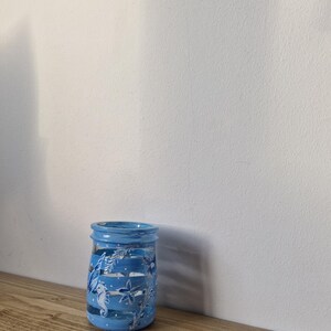 May include: A blue painted glass jar with a coastal theme. The jar features white seahorses, starfish, and floral designs. The jar is sitting on a wooden surface.