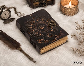 May include: A black leather-bound journal featuring a gold-toned embossed eye, sunburst, moon, and cloud design on the cover. The journal includes a metal latch closure and thick, cream-colored pages with deckled edges, accompanied by a quill pen.