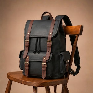 Può includere: Uno zaino nero con cinghie marroni effetto pelle e fibbie in metallo color ottone. La borsa presenta una patta superiore, una tasca frontale e due tasche laterali, tutte chiuse con fibbie regolabili e un'apertura con coulisse.