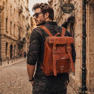 May include: A brown leather backpack with adjustable straps and buckle fastenings. The backpack has a rectangular shape with a flap top and a front pocket. The person is wearing sunglasses and a black shirt.