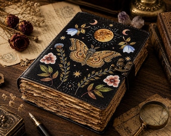 May include: A black journal with a metal latch featuring a moth, a full moon, stars, and floral illustrations on the cover. The book has thick, deckled-edge pages that appear aged and textured, creating a distinct look for this bound notebook.