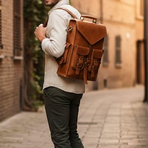 Puede incluir: Una mochila de cuero color canela con asa superior, solapa con cierre de hebilla y dos bolsillos frontales con correas de hebilla. La bolsa cuenta con correas ajustables para los hombros y herrajes metálicos laterales de diseño estructurado.