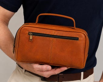 Peut inclure: Une trousse de toilette rectangulaire de couleur fauve avec une poignée supérieure et une poche zippée horizontale sur le devant. Le sac présente une texture lisse semblable à du cuir, des coutures visibles et une tirette de fermeture éclair en métal.