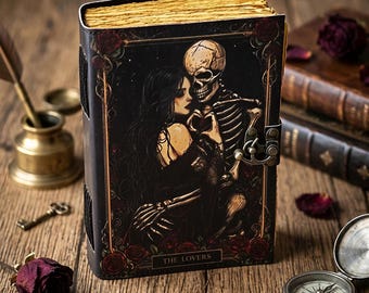 May include: A rectangular journal with a dark cover featuring an illustration of a person and a skeleton embracing, labeled THE LOVERS. The book has gold-toned edges, a metal latch closure, and decorative red rose borders on the front cover.
