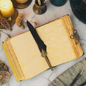May include: A rectangular journal with thick, cream-colored pages and a metal latch closure, featuring a black feather quill pen with a gold-toned leaf-shaped metal accent resting diagonally across the open pages.