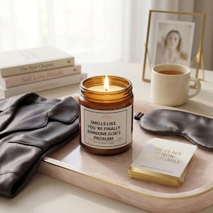 May include: A lit soy candle in a brown glass jar with the text "Smells like you're finally someone else's problem." The candle sits on a pink tray with a sleep mask, a stack of books, and a cup of tea. A framed photo is also on the tray.