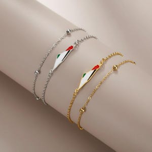 May include: Two bracelets, one silver and one gold, each featuring a diamond-shaped charm with a red, green, and white design. The bracelets have delicate chain links and small ball accents.