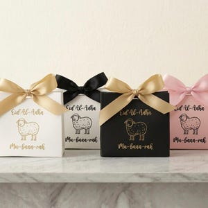 Puede incluir: Cuatro cajas de regalo con lazos dorados, negros y rosas. Cada caja tiene una ilustración dorada de una oveja y las palabras "Eid Al-Adha" y "Mu-baaa-rak" en letras doradas. Las cajas son blancas, negras y rosas.