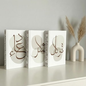 Pode incluir: Três livros de capa dura brancos dispostos verticalmente, cada um apresentando caligrafia árabe castanha centrada sobre formas circulares bege suaves. As lombadas contêm os textos Alhamdulillah, Allahu Akbar e Subhanallah em preto.