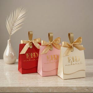 Puede incluir: Tres bolsas de regalo en rojo, rosa y crema, cada una adornada con una cinta y asa doradas. Las bolsas presentan el texto dorado "EID mubarak". Un jarrón blanco con una hoja decorativa está en el fondo.