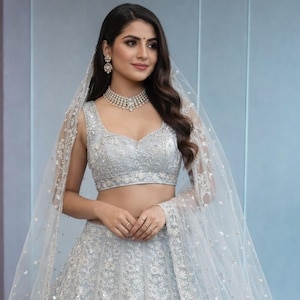 May include: A light blue, sleeveless, embellished lehenga with a matching crop top and a sheer veil. The outfit features intricate embroidery and is accessorized with a diamond necklace and earrings. The woman is wearing a light blue outfit.