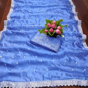 Könnte beinhalten: Ein blaues Seidensari mit einem passenden Blusenteil. Das Sari hat einen silbernen, gewellten Rand und verstreute silberne Verzierungen. Ein Strauß rosa Blumen liegt auf dem Sari. Der Text "SILKSTOREIndia" ist sichtbar.