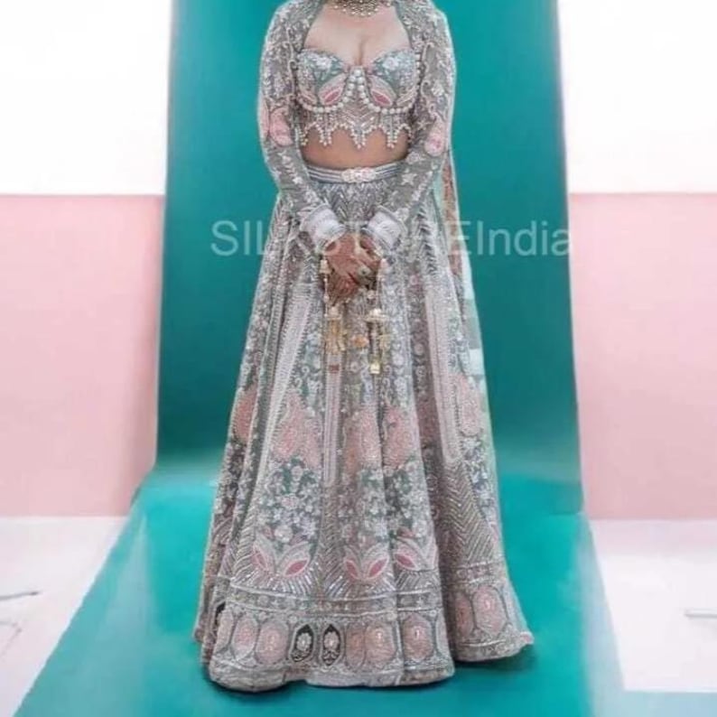 May include: A long-sleeved lehenga set featuring a cropped bodice and a floor-length skirt in shades of sage green and soft pink. The garment is heavily embellished with intricate silver embroidery, beadwork, and sequins throughout the fabric.