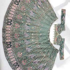 May include: A sage green lehenga skirt and matching blouse set featuring intricate embroidery in pink, white, and metallic gold thread. The skirt displays a wide border with repeating oval motifs, while the blouse has a scalloped hemline.