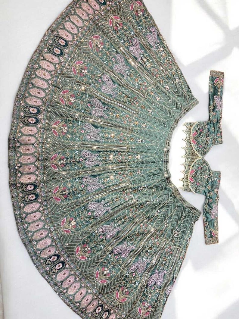 May include: A sage green lehenga skirt and matching blouse set featuring intricate embroidery in pink, white, and metallic gold thread. The skirt displays a wide border with repeating oval motifs, while the blouse has a scalloped hemline.