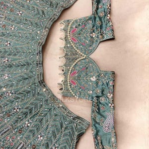 May include: A sage green lehenga set featuring a long-sleeved blouse and a flared skirt. Both pieces are adorned with intricate silver embroidery, small mirrors, and pink floral accents. The blouse neckline is trimmed with a row of white pearls.
