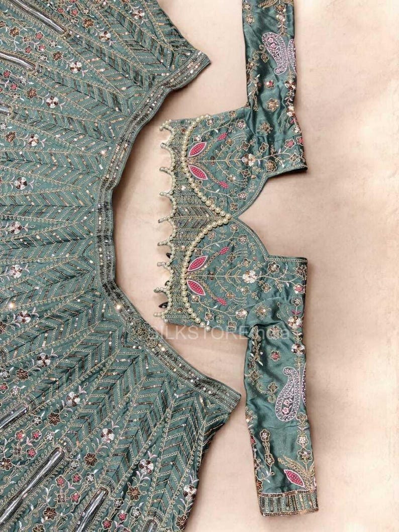 May include: A sage green lehenga set featuring a long-sleeved blouse and a flared skirt. Both pieces are adorned with intricate silver embroidery, small mirrors, and pink floral accents. The blouse neckline is trimmed with a row of white pearls.