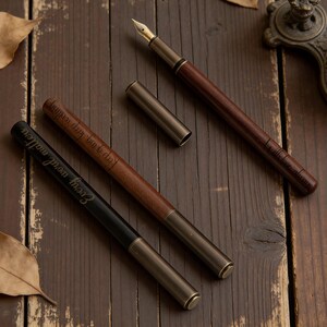 May include: Three fountain pens with brown and black barrels and gold-colored nibs and accents are displayed on a wooden surface. One pen has the text "Every word matters" engraved on it. A pen holder is in the background.