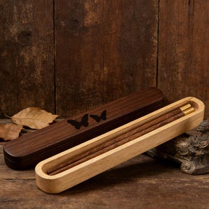 May include: Wooden chopsticks with gold accents rest in a light-colored wooden holder. A dark brown wooden case with butterfly silhouettes is next to the holder. The chopsticks and case are on a wooden surface.