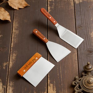 May include: Three stainless steel grilling spatulas with wooden handles. The handles have the phrases "Food is love", "Sizzle & serve", and "Grill with joy" engraved on them. The spatulas are on a dark brown wooden surface.