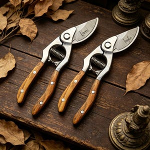 May include: Two gardening shears with wooden handles and silver blades, each engraved with a name. The shears are set on a dark wooden surface, surrounded by dried leaves and decorative objects.