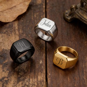 May include: Three signet rings in silver, black, and gold, each with a personalized engraving. The silver ring is engraved with the name "Julia", the black ring with the letter "M", and the gold ring with the letter "B". The rings are displayed on a wooden surface.