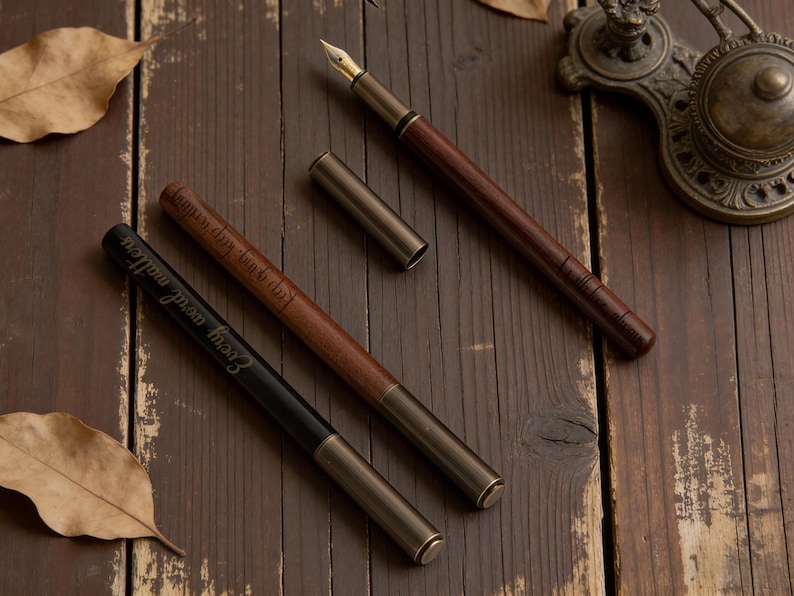 May include: Three fountain pens with brown and black barrels and gold-colored nibs and accents are displayed on a wooden surface. One pen has the text "Every word matters" engraved on it. A pen holder is in the background.