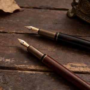 May include: Two fountain pens with gold nibs and dark wooden barrels rest on a weathered wooden surface. One pen is a deep brown, the other is black. Dried leaves and a decorative metal piece are in the background.