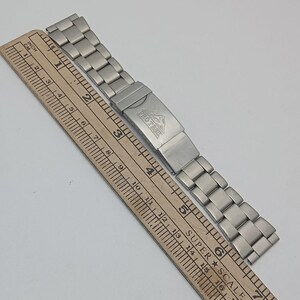 May include: A silver-toned watch band with a folding clasp, featuring the text "PRO TREK" and a mountain logo. A wooden ruler with inch and centimeter markings is next to the watch band.