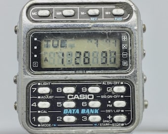 May include: A vintage Casio Data Bank digital watch with a silver-tone metal band and case. The watch face displays the time and date in a digital format. The watch has multiple buttons and functions, including light, adjust, and mode.